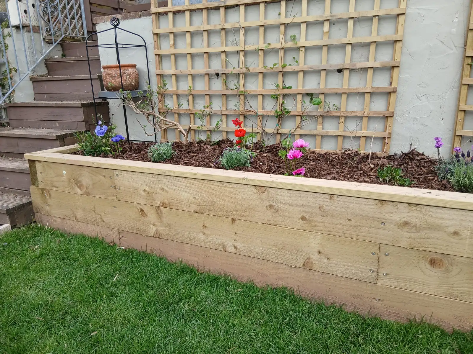 Pressure-treated timber sleeper raised flower bed topped with bark mulch, beside a lawn with square timber trellis behind